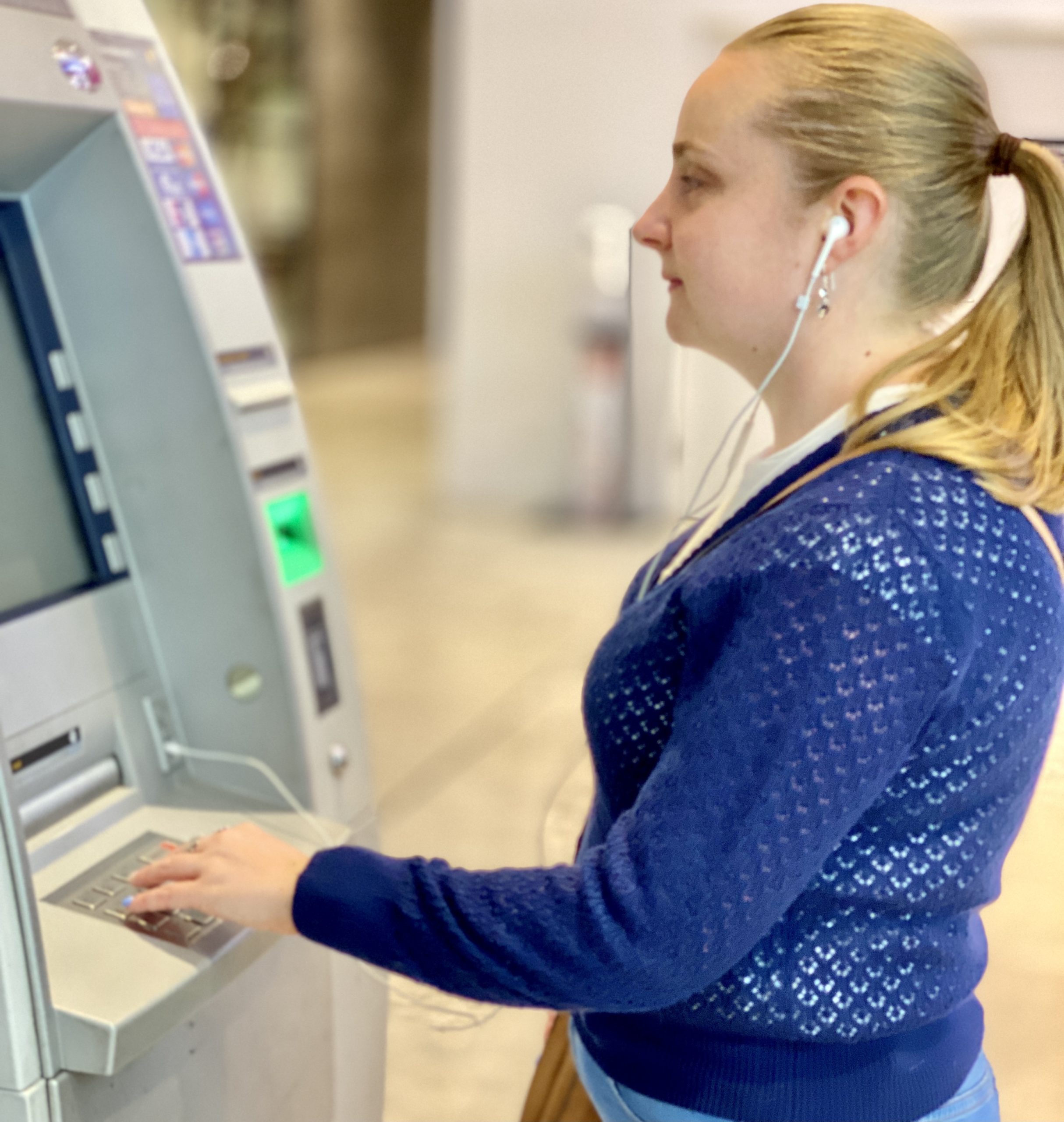 Cass stands in front of an ATM with her hand on the keypad. She is wearing a royal blue lace-knit cardigan with a white short-sleeve blouse underneath and light blue jeans.