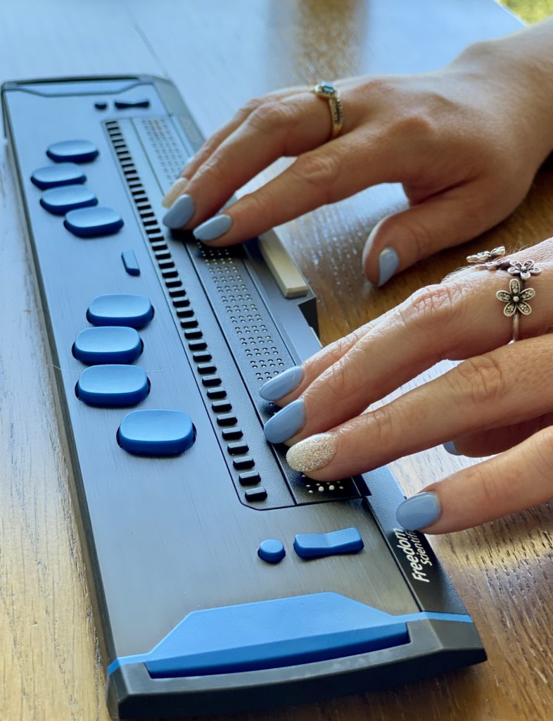 Cass’s hands rest lightly on a black braille display which sits on a table. Small black keys sit in front of her hands on the display, with larger blue buttons lining the front of the display. In the bottom left of the display reads “Freedom Scientific” in white text. Her nails are painted a light blue, with her ring fingers painted a sparkly white. She wears a silver flower ring on her left hand and a gold ring with a blue gem on the right. Photo by Lee Anderson.