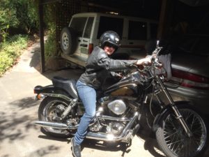 Cass, a young woman is sitting on a Harley motorbike. She is smiling and it is a lovely sunny day. She is wearing a kick-ass biker jacket, jeans and black shiny helmet, and she looks cool as. Behind her is a parked car and lush greenery.