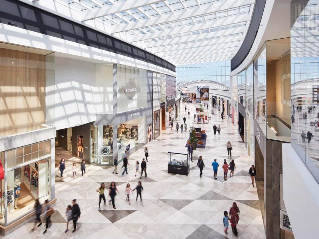 Beautifully lit photograph from a higher angle of the inside of Pacific Werribee shopping centre- glass diamond shaped ceiling panels and marble mozaic floor with people walking around shopping.