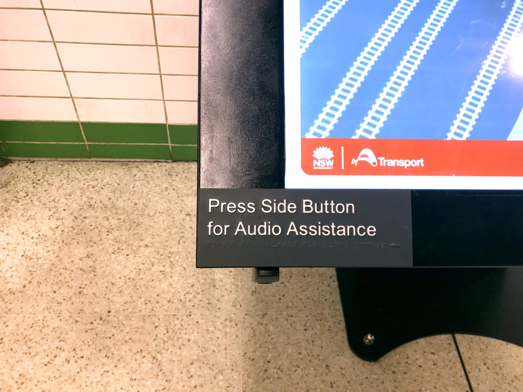 Image of an information kiosk screen with the words 'Press side button for audio assistance' written in english and then there is braille underneath
