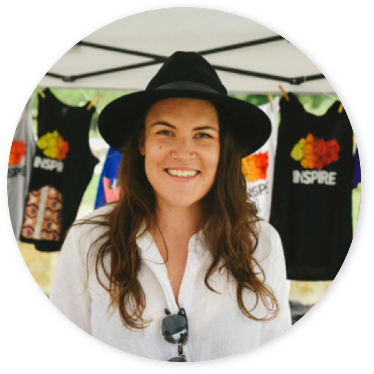 Photograph of a smiling woman wearing a wide brimmed black hat, with her long brunette hair flowing out over her white blouse. Just behind her is a market stall with tshirts hanging up.